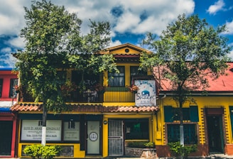 A vibrant building with a yellow facade and a red roof, surrounded by leafy trees and blue skies. The structure has a sign for a Shakti establishment and another sign for a lawyer's office. The windows are framed with green shutters, and there are several plants in front of the building.