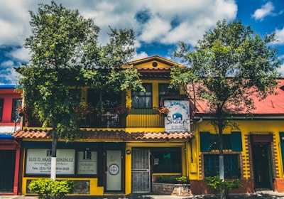 A vibrant building with a yellow facade and a red roof, surrounded by leafy trees and blue skies. The structure has a sign for a Shakti establishment and another sign for a lawyer's office. The windows are framed with green shutters, and there are several plants in front of the building.